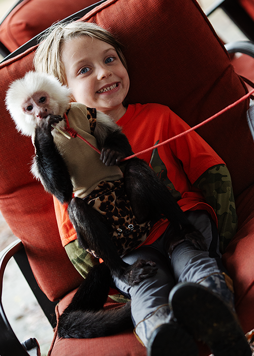 A young girl sitting on an orange chair, holding a black monkey wearing a beige vest with leopard print trim, in an indoor setting.