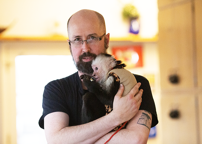 A man with glasses and a beard holding a baby skunk indoors.