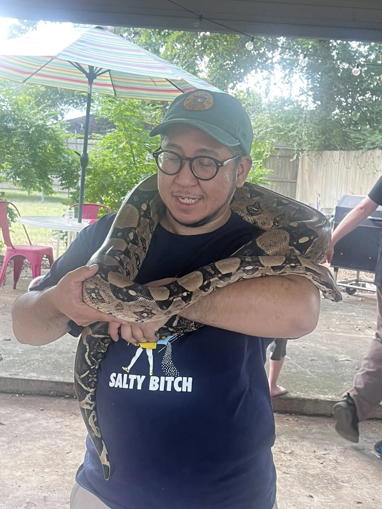 A man with glasses, wearing a baseball cap, smiles with closed eyes as a snake drapes over his shoulders. The man is wearing a black T-shirt with the words 'Salty Bitch' partially visible. The background shows a backyard with trees, a patio umbrella,