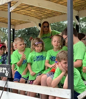 Group of children and a woman sitting on a parade float or tiered platform, all wearing matching green shirts, with some children clapping and others looking around, outdoors with trees in the background.