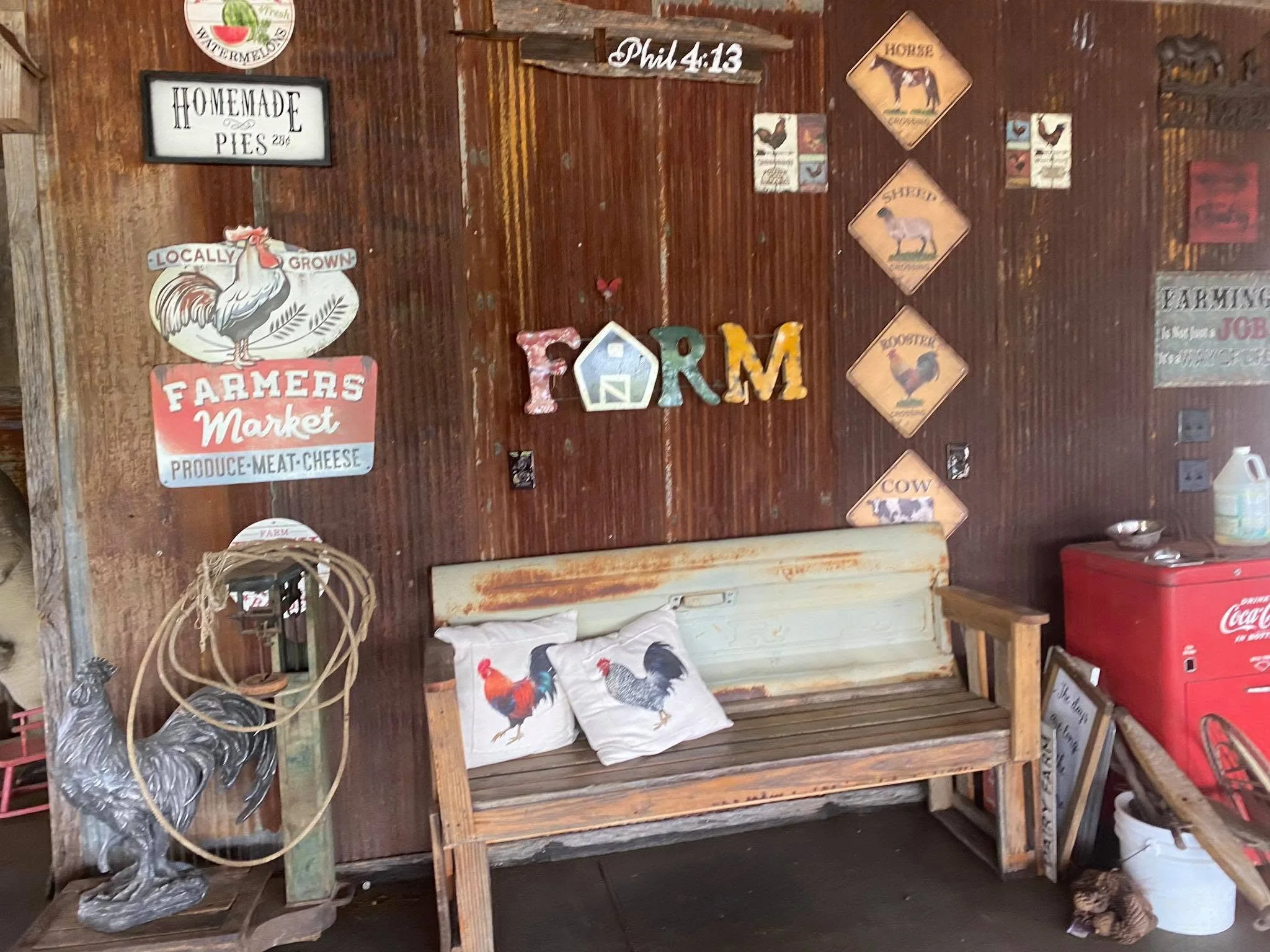 A rustic wooden bench decorated with chicken-themed pillows and surrounded by various farm-inspired signs and ornaments, including a rooster sign and vintage metal farm tools.
