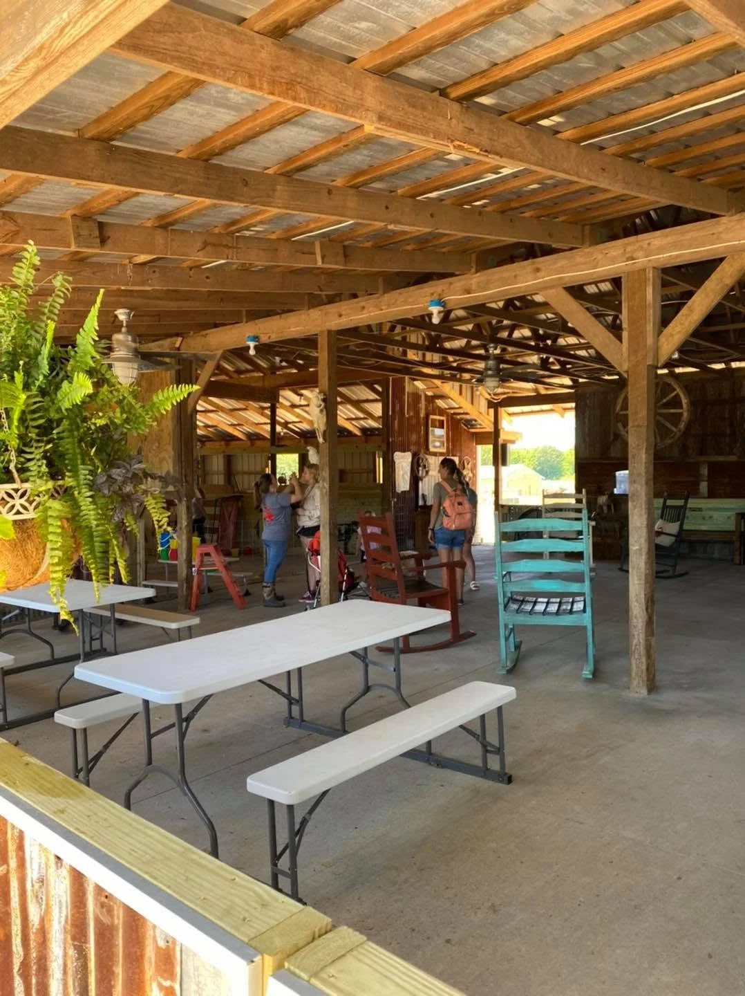 Indoor rustic barn with wooden rafters, ceiling fans, and outdoor style furniture including picnic tables, rocking chairs, and upside-down chairs, with a group of people gathered inside.