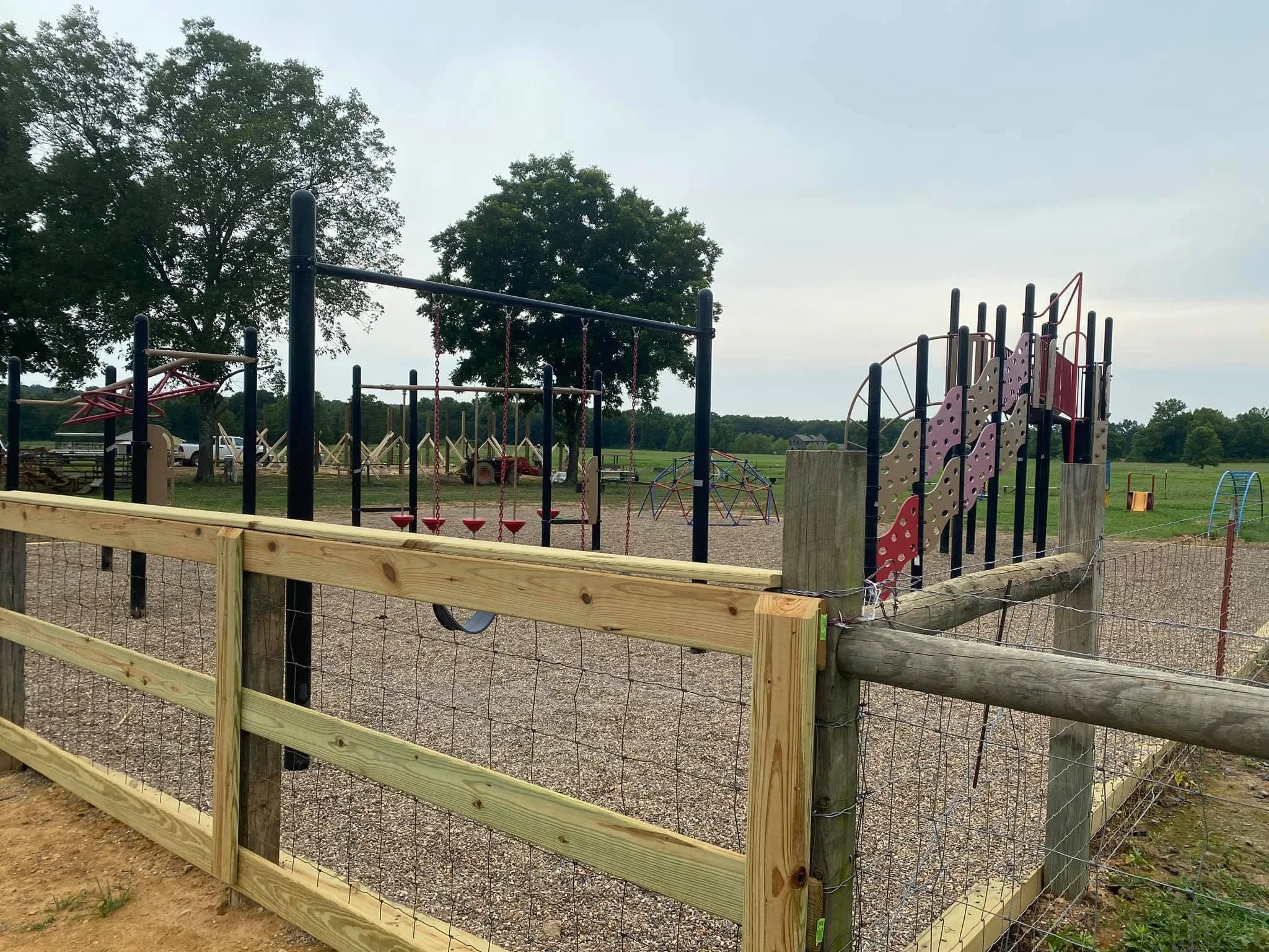 Playground with swing set, climbing structures, and safety fencing, situated in a rural area with trees and open fields in the background.