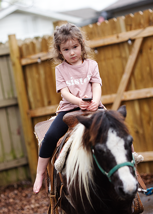 Young girl with curly hair riding a small horse or pony, holding a phone, in a backyard with a wooden fence.