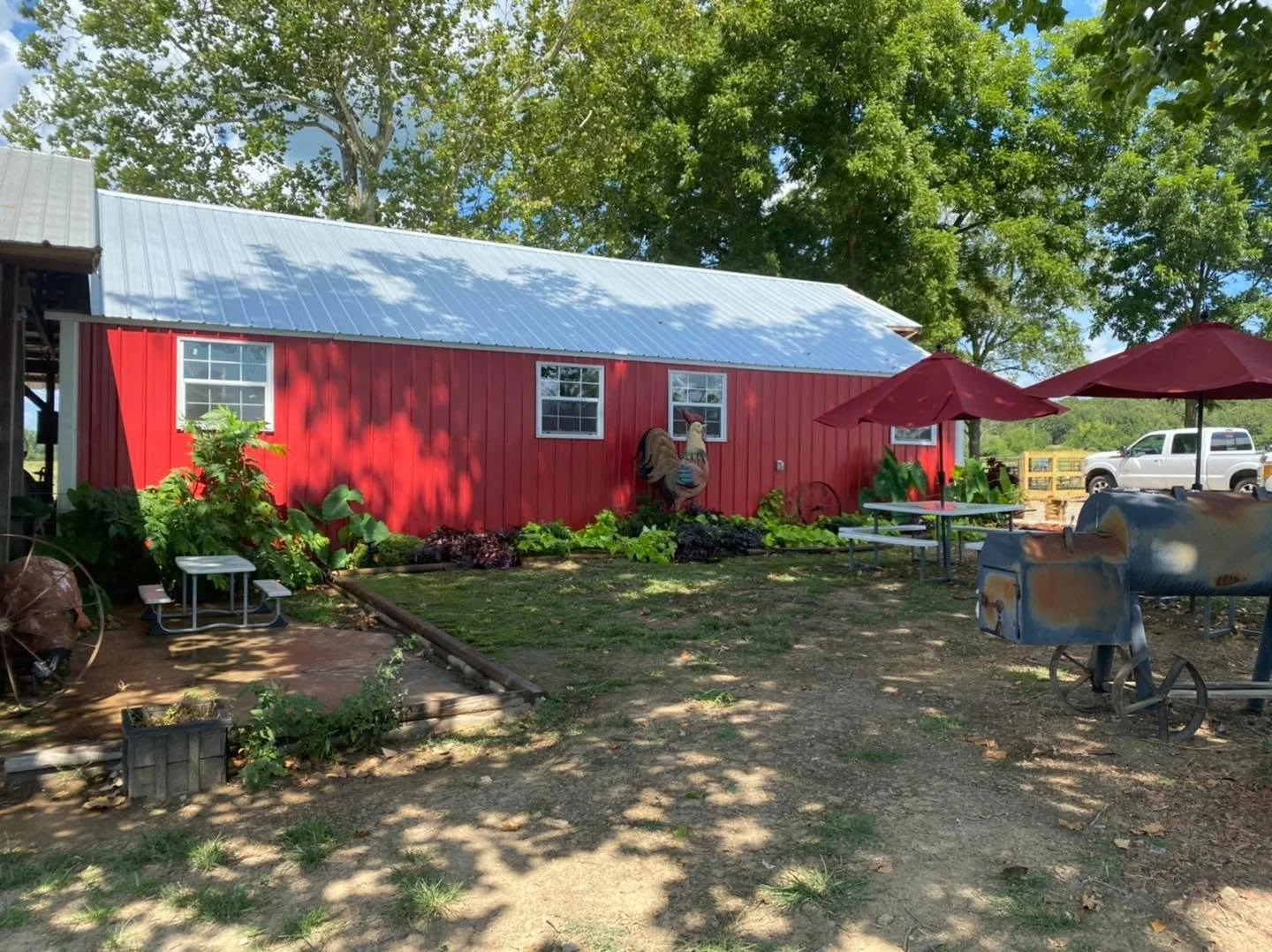 Red barn with white windows, garden with green plants, decorated with a rooster sculpture, outdoor seating with red umbrellas, and a vintage barbecue smoker in front.
