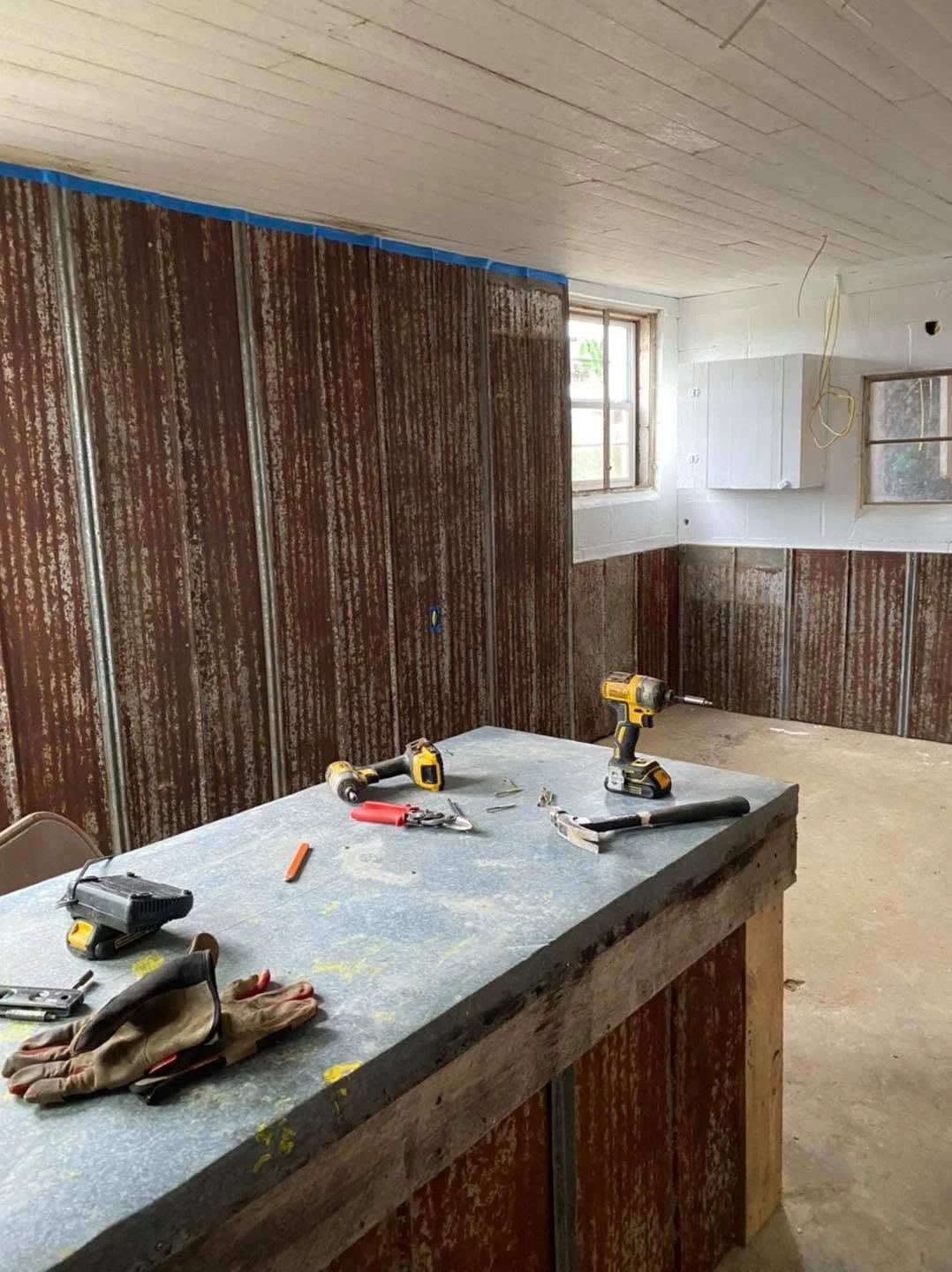 Interior of a room under renovation with tools on a worktable, metal wall panels being installed, and two windows providing natural light.
