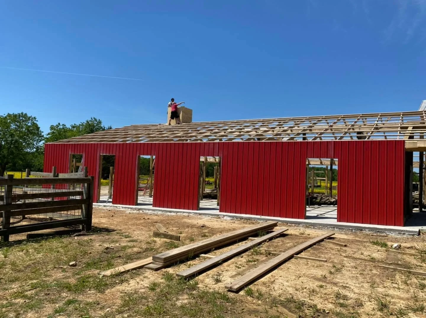 Construction site with a building in progress, red exterior walls, partially completed roof framework, and a worker standing on the roof.