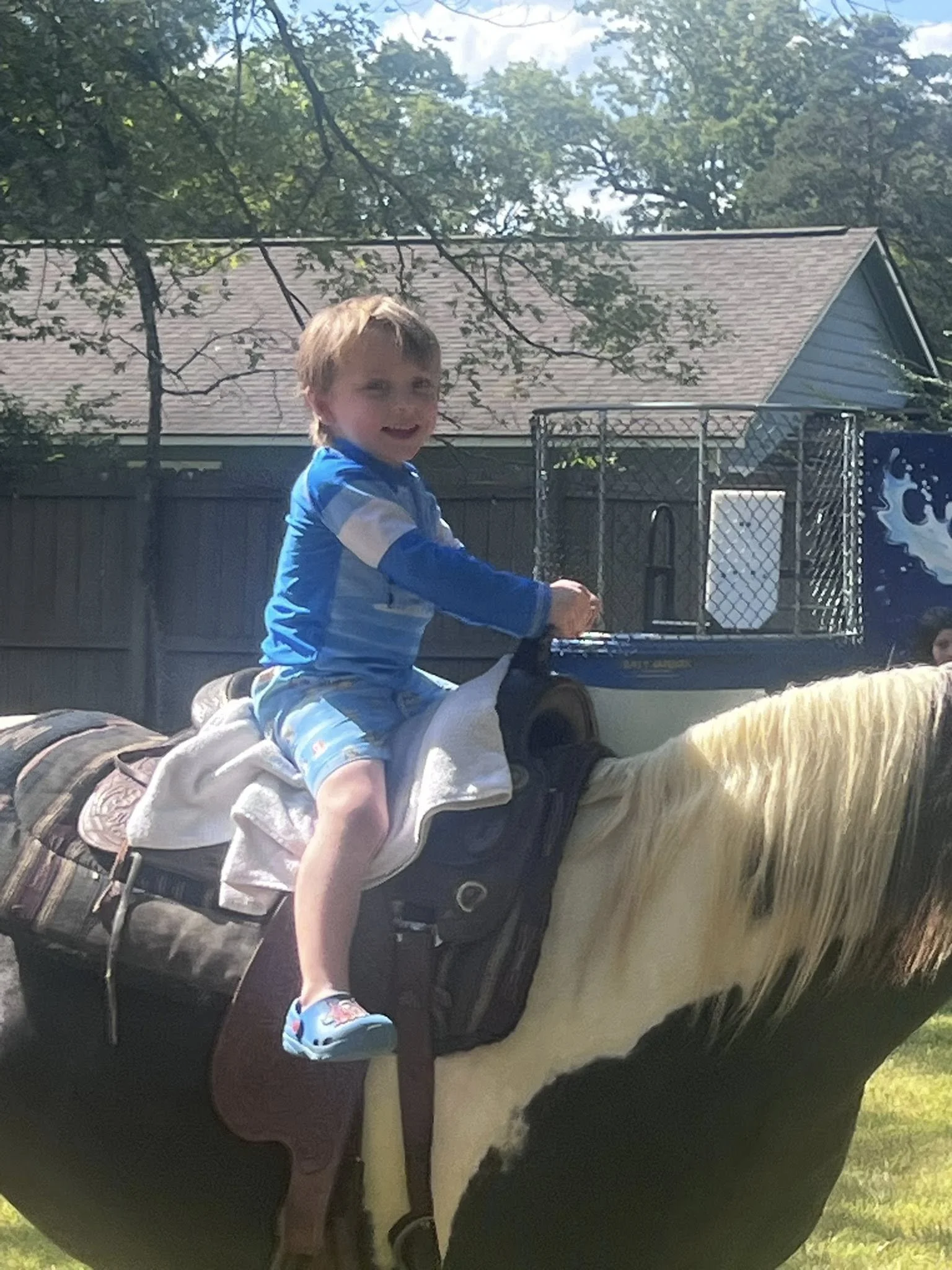 A young boy with light brown hair smiling while riding a black and white horse outdoors. The boy is wearing a blue and white jacket, blue shoes with cartoon characters, and is holding onto the horse's saddle. The background shows a fence, trees, and 