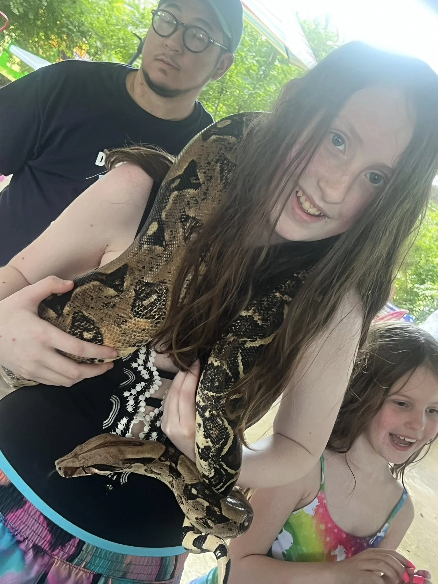 A young girl and a man with glasses and a cap share a moment at a beach. The girl has wet hair, a pink tie-dye swimsuit, and a wide smile. The girl is holding a snake with brown and black markings on her arm. The background shows green trees and a co