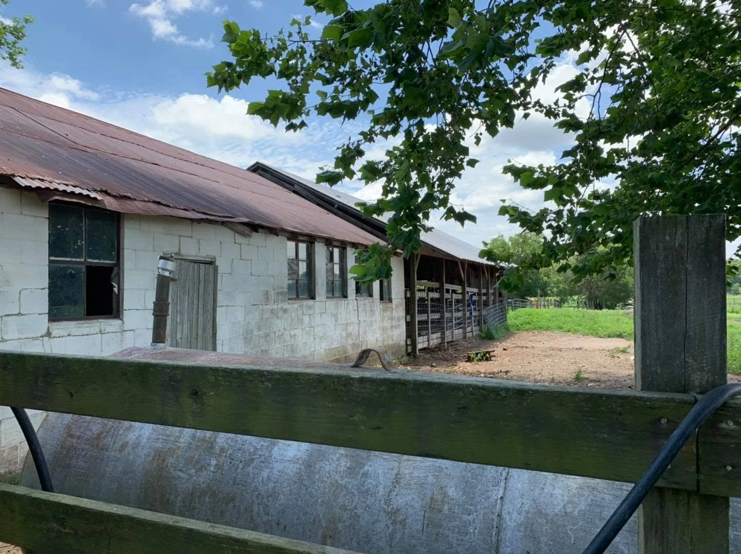 A rustic farm building made of white cinder blocks with a rusted metal roof. There are several small windows and a closed wooden door. A wooden fence is in the foreground, and a tree with green leaves partially frames the scene. The sky is partly clo