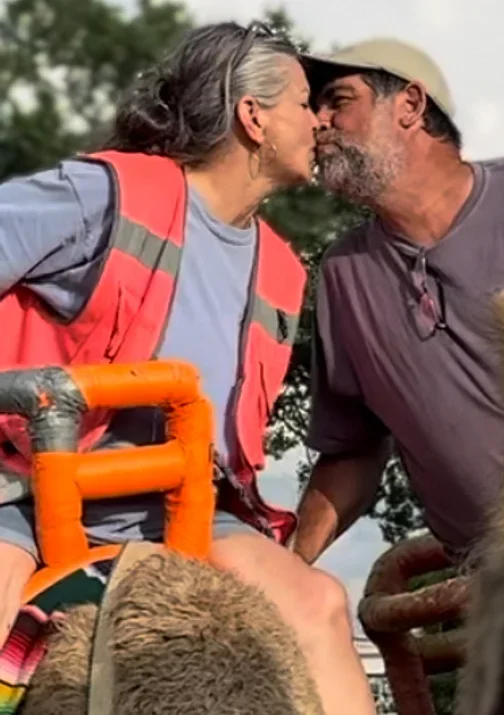 An elderly woman and a man are sharing a kiss outdoors, with trees and a cloudy sky in the background. The woman is sitting in a wheelchair, wearing a gray shirt and a red vest, and the man is leaning in toward her, wearing a gray shirt and a cap.