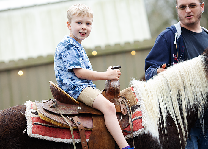 A young boy riding a small horse with a saddle, holding the reins, and smiling. An adult is standing beside the horse, holding the reins and guiding the boy.