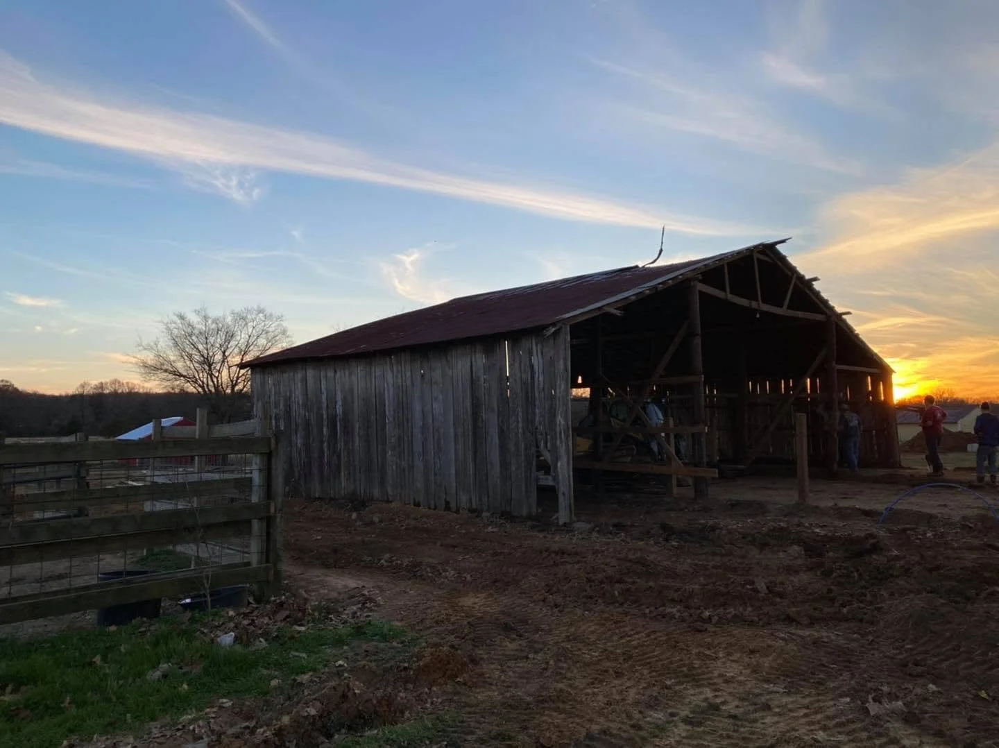 A rustic wooden barn under construction at sunset, with a dirt ground and a few people working nearby, on a rural farm landscape.