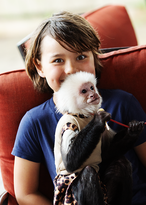 Young girl with short brown hair smiling while holding a baby capuchin monkey on her lap, sitting on a red couch.