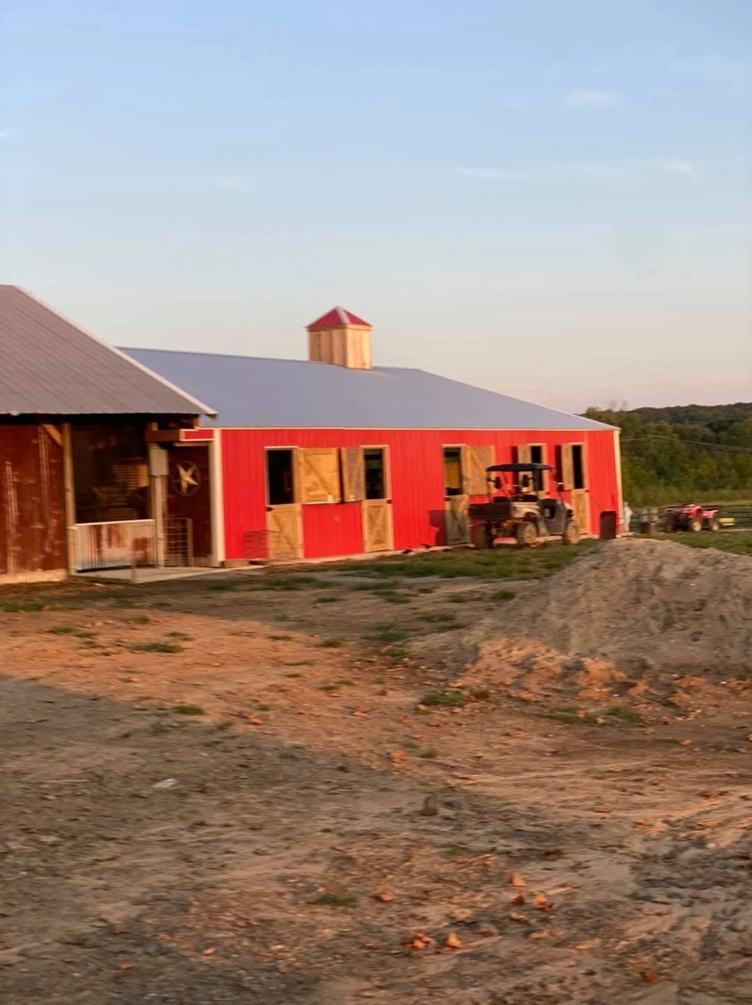 This image shows a farm building with a red exterior and a gray roof, some vehicles including a tractor, and a landscape with green fields and distant trees under a clear sky.