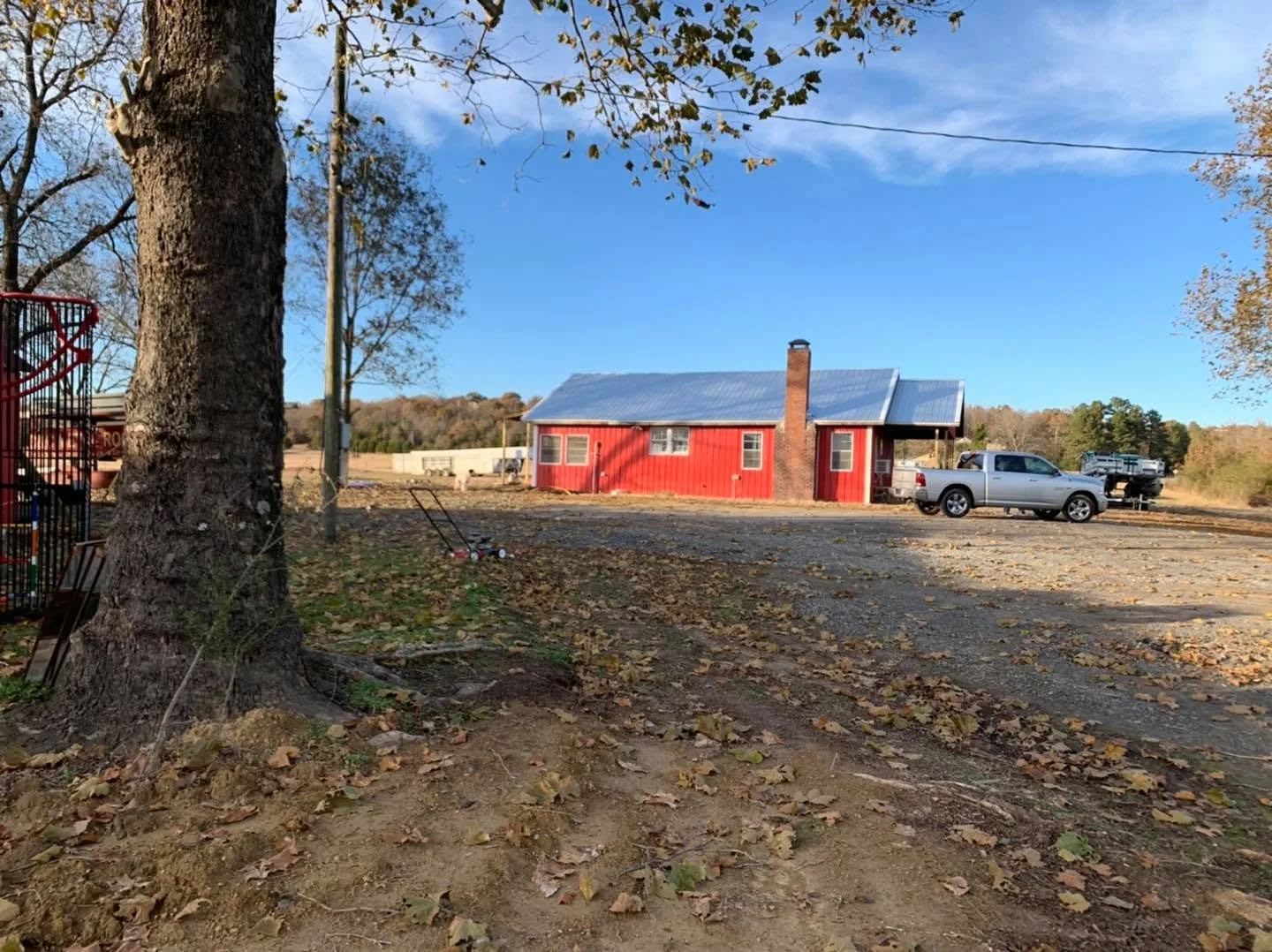 A rural scene with a large tree in the foreground, a red building with a metal roof, a gray pickup truck, and a landscape with a clear blue sky and some scattered clouds.