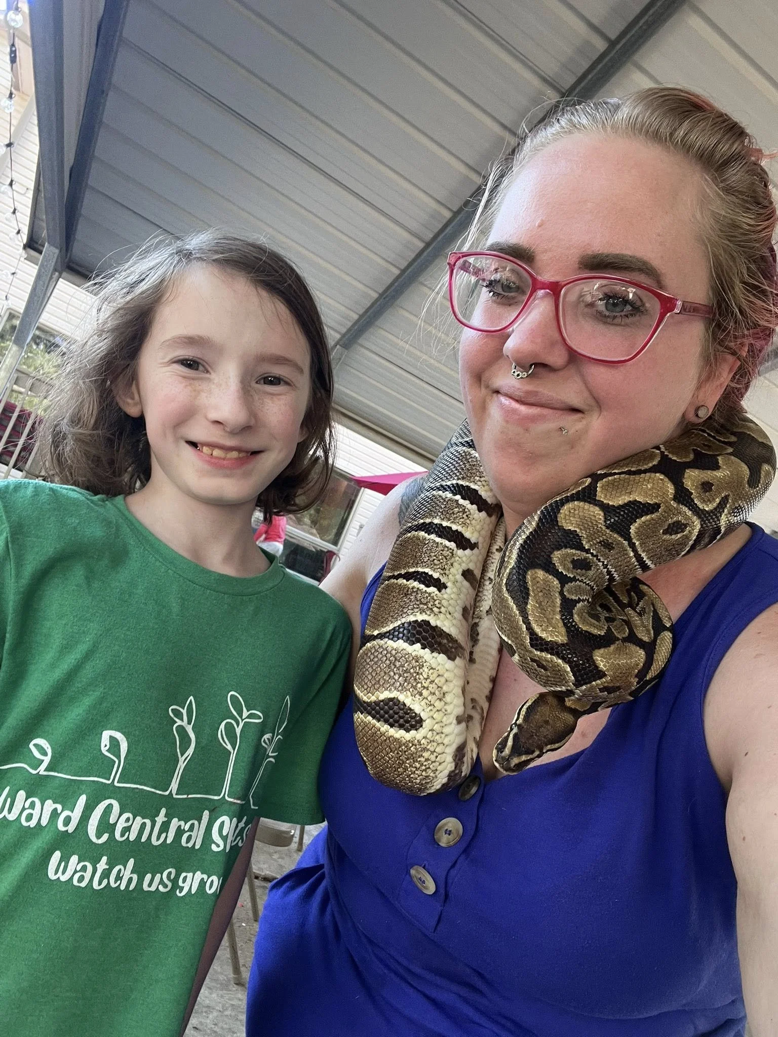A woman with glasses, a septum piercing, and a snake around her neck smiling at the camera. A young girl with brown hair and freckles, wearing a green shirt, also smiling. They are under a metal roof with a building and outdoor setting in the backgro