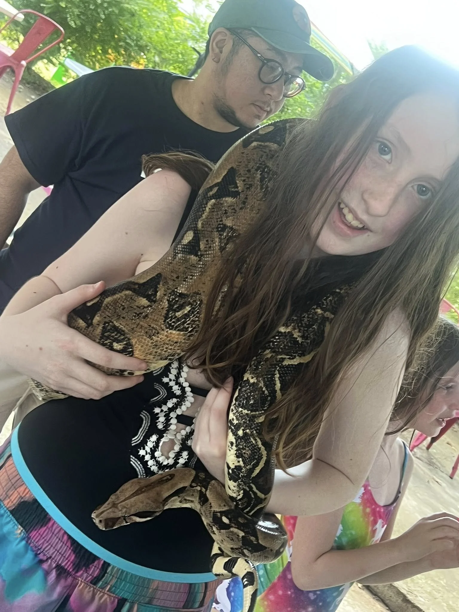 A young woman with red hair and freckles holding a large snake on her arm, at a festival with people in colorful swimsuits and tropical shirts.
