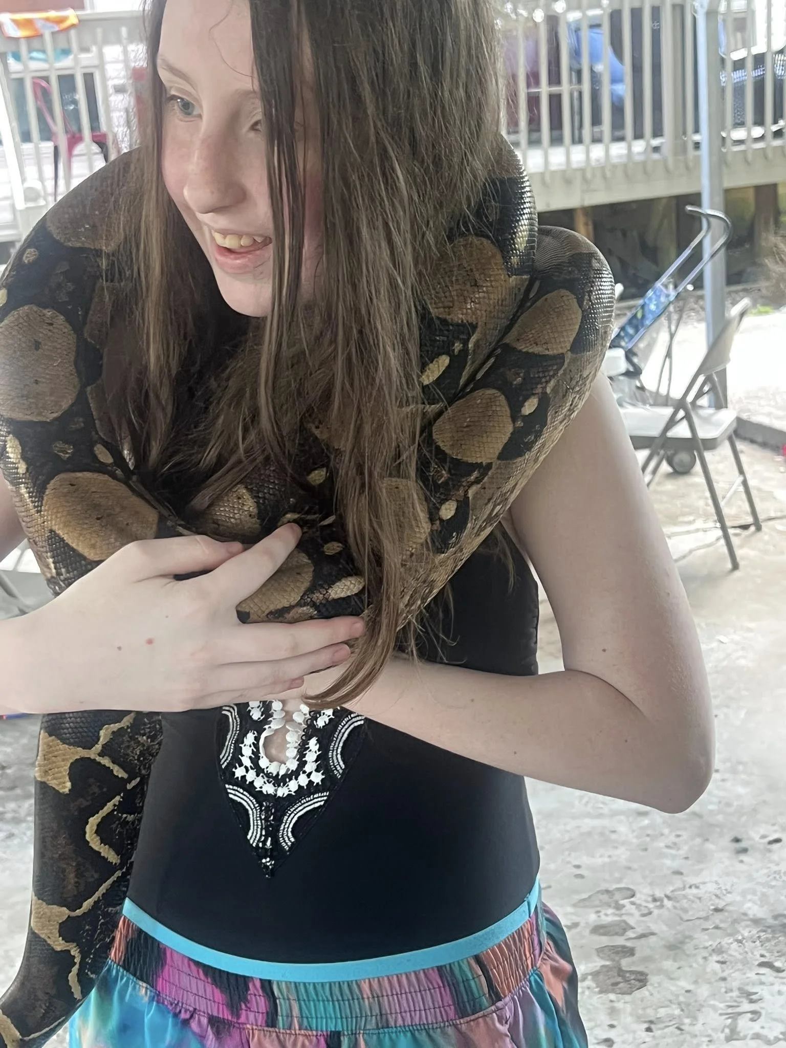 A young girl with long brown hair and blue eyes, smiling with her mouth open, showing her teeth. She is wearing a black top with white embroidery and multicolored shorts. She is holding a large snake around her shoulders and arm. The background inclu