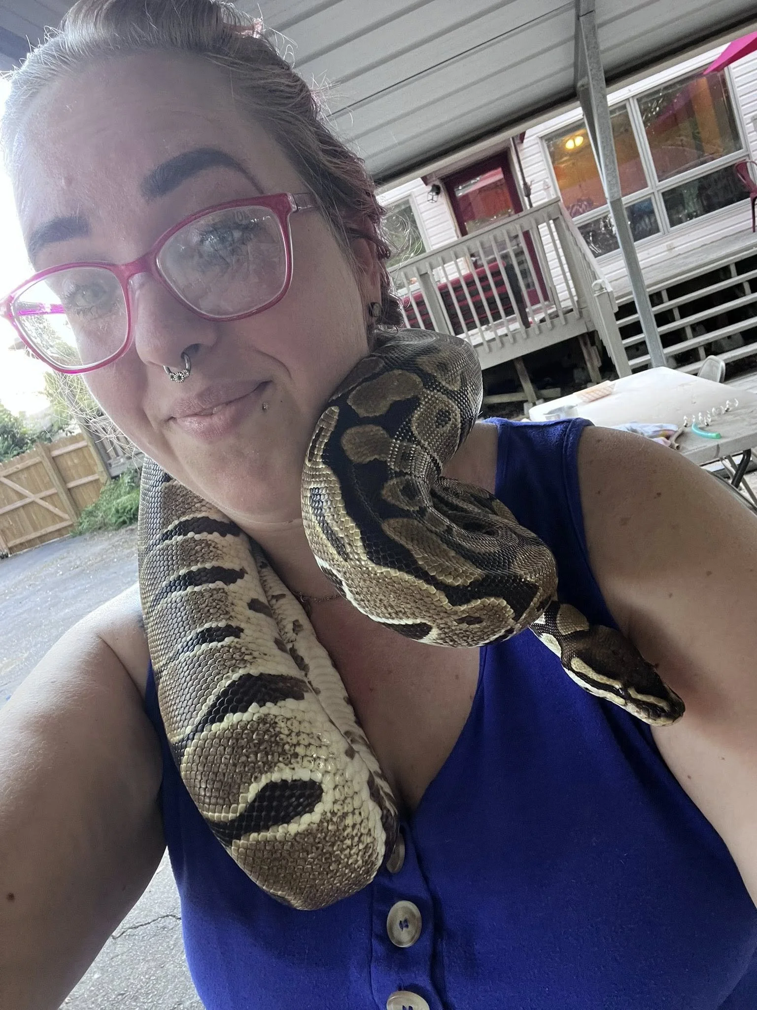 A woman with glasses and multiple facial piercings holding a pet ball python snake around her neck outside a house with a porch and stairs.
