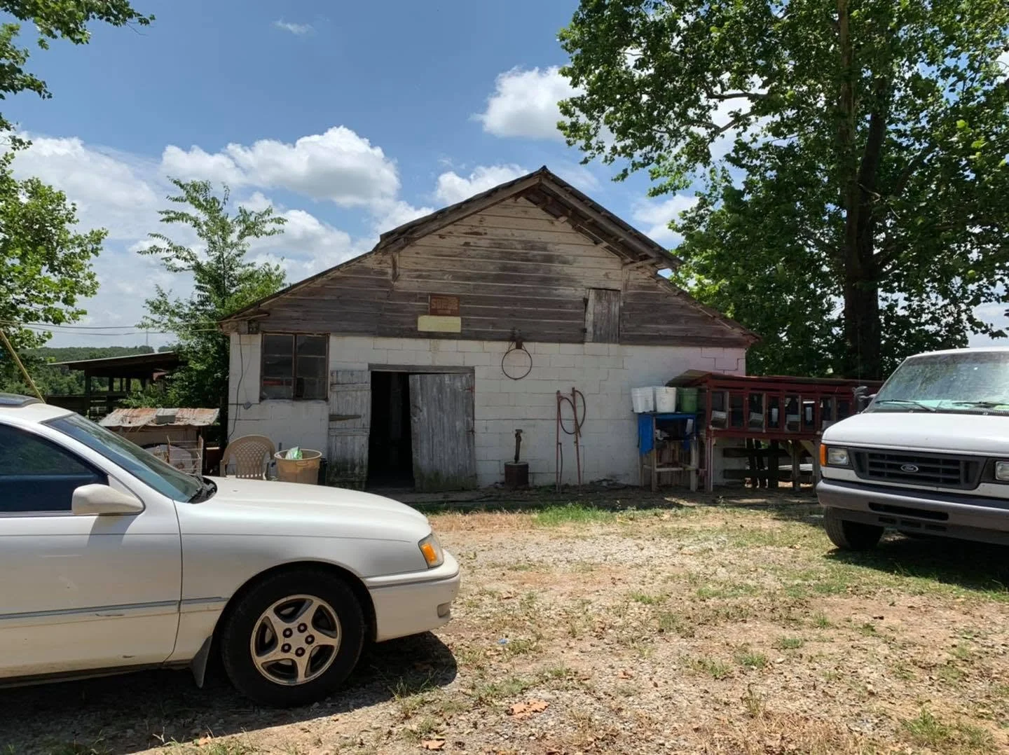 An old rustic barn with a weathered wooden upper section and white brick lower section, with two cars parked in front under a bright, partly cloudy sky, surrounded by green trees.