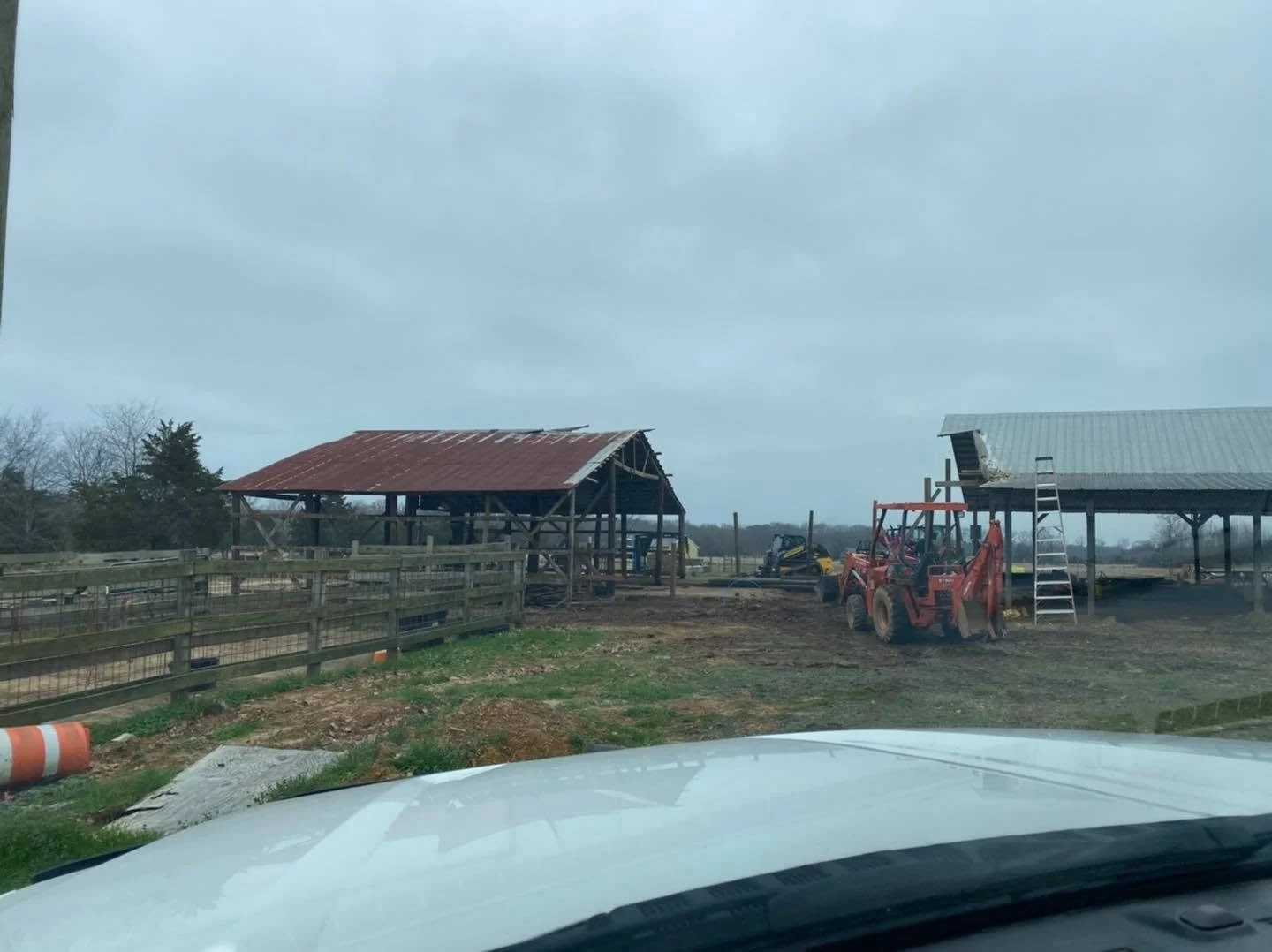 A farm scene with two partially built barn structures, construction equipment, and fencing, seen through a vehicle windshield on a cloudy day.