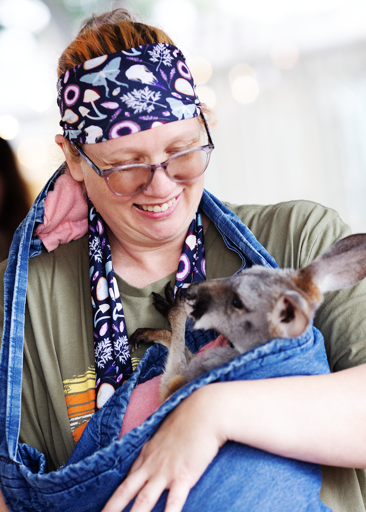 A person with red hair, glasses, and a colorful headband is holding a small dog wrapped in a blue blanket, smiling down at it.