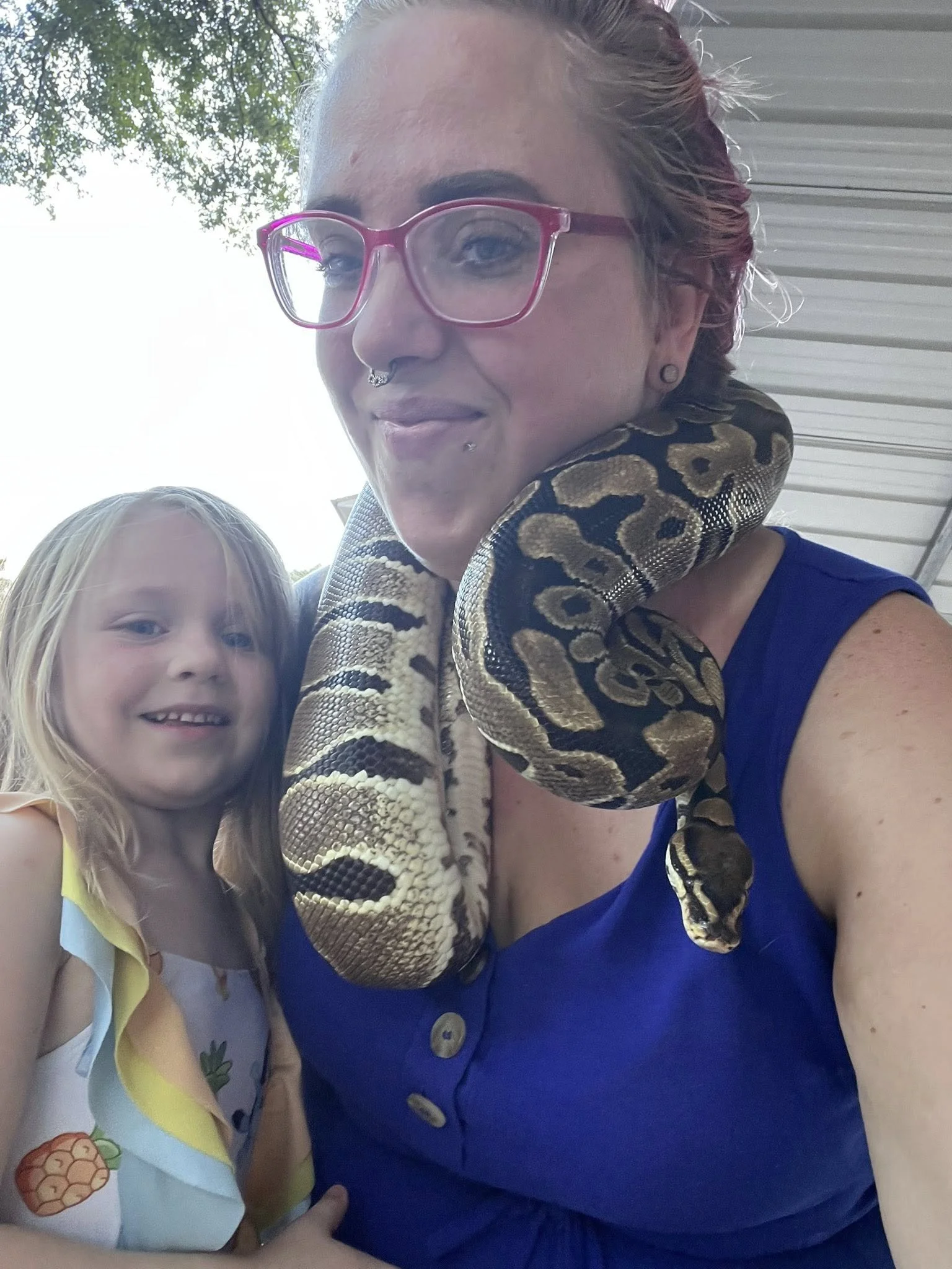 A woman with short red hair, pink glasses, and multiple facial piercings, holding a snake around her neck, standing next to a young girl with long blonde hair and a pineapple print dress, outdoors with trees and a building in the background.