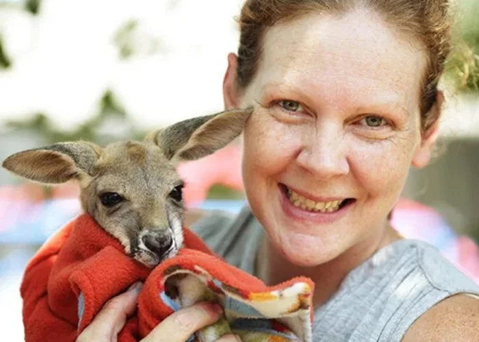 A woman smiling and holding a small dog wrapped in a red blanket outdoors.