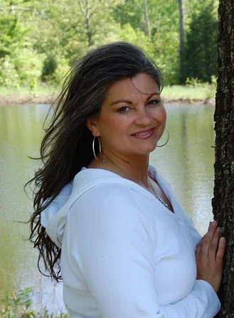 A woman with long dark hair wearing a white hoodie, standing outdoors near a tree, with a lake and green trees in the background.
