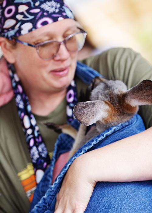 Person holding a small dog wrapped in a blanket, wearing glasses and a colorful headscarf.