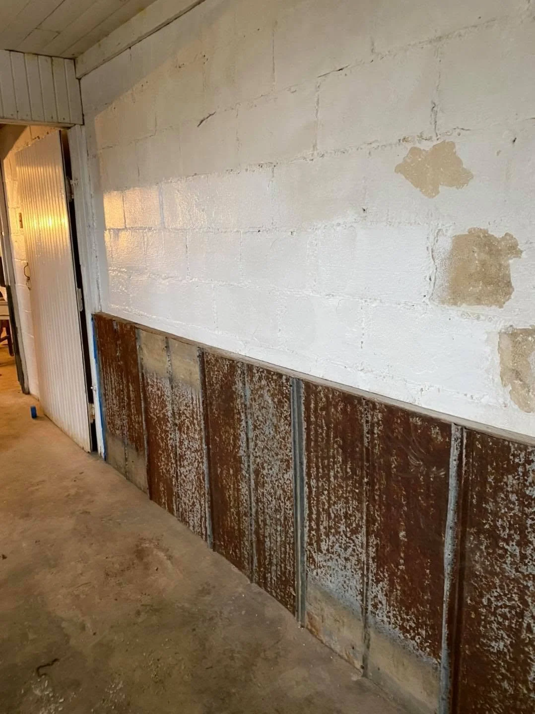 Interior wall with the lower half covered in rusted steel panels and the upper half painted white, showing patches and damage, in a possibly unfinished room.