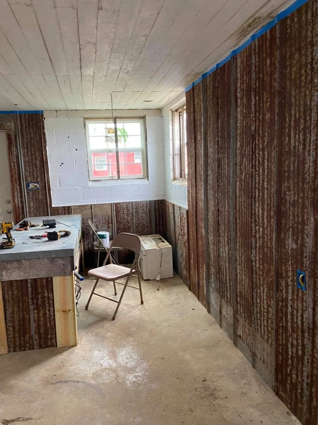 Room under renovation with exposed walls and ceiling, construction tools on a work table, a folding chair, and uninstalled electrical outlets, with two windows near the ceiling.