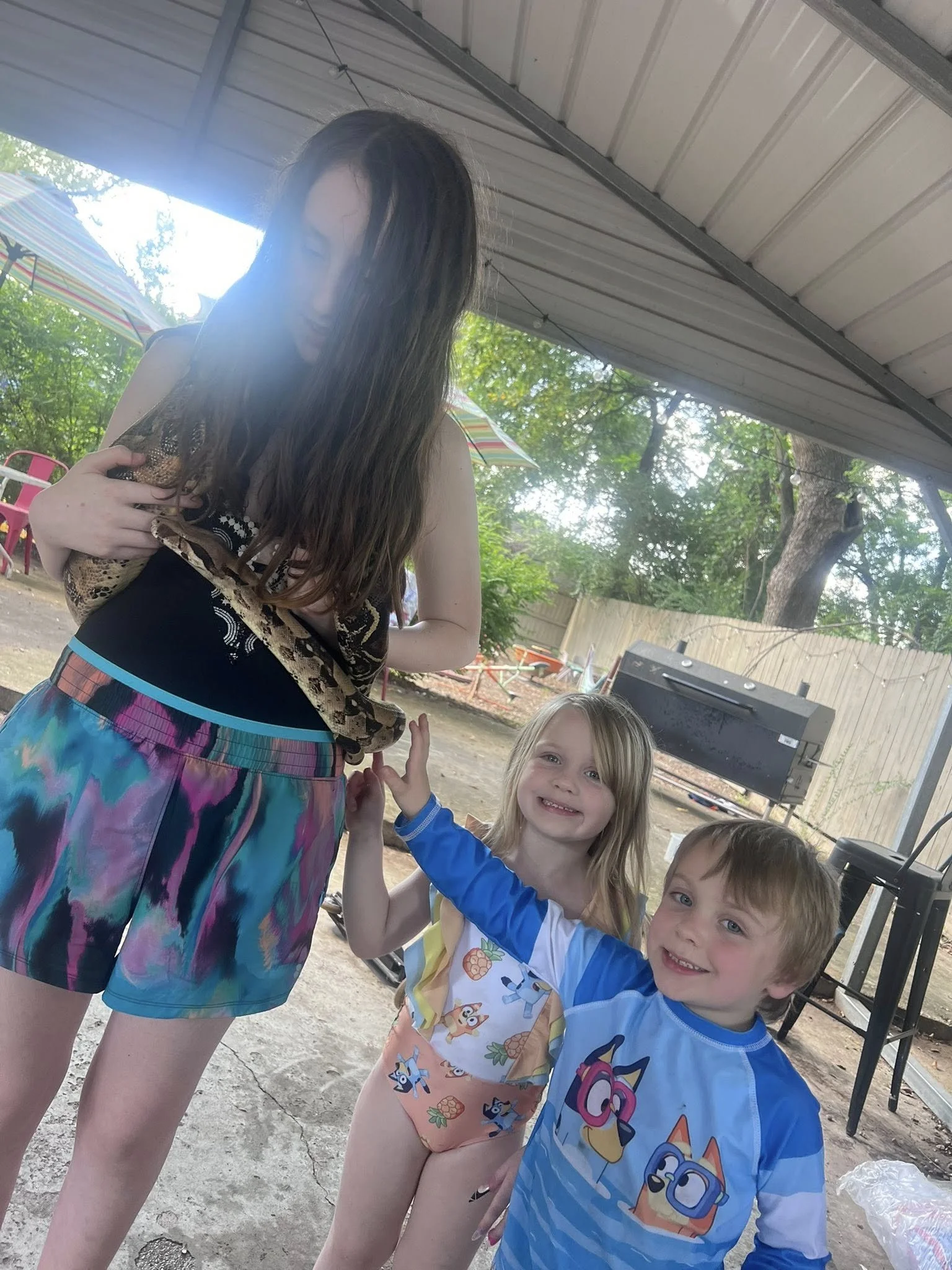 A person holding a snake with three children smiling and giving a high five outdoors in a backyard with a fence, trees, and colorful umbrellas.
