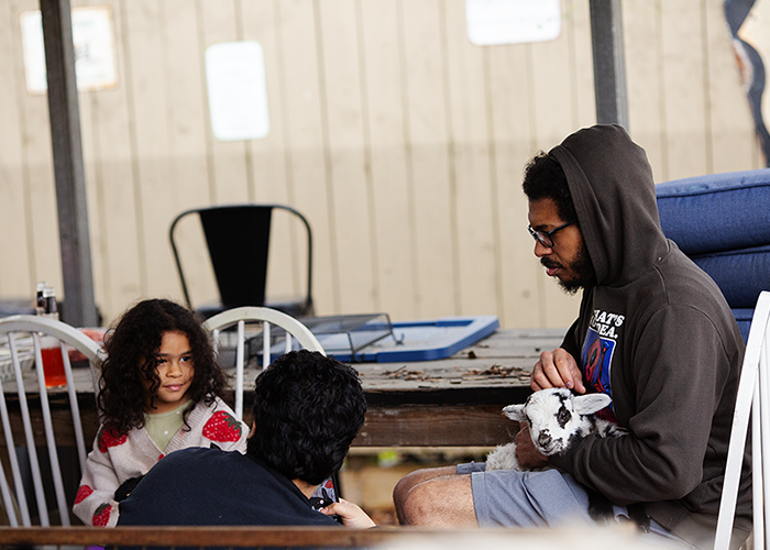 A man with glasses in a hoodie sitting on a white chair holding a guinea pig, while a young girl in a white sweater with red strawberries watches and a person with dark hair in a dark shirt faces away from the camera in a rustic room.