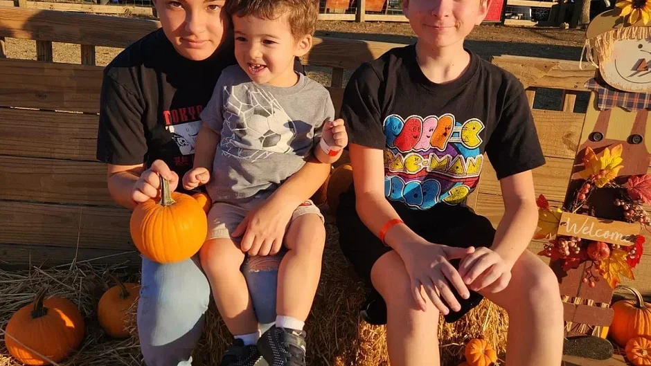 Three kids sitting on hay bales outdoors surrounded by pumpkins, with fall decorations and a sign that says "Welcome."