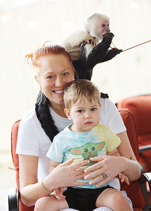 A smiling woman with red hair holding a young boy with blond hair on her lap, with a monkey on her shoulder. The monkey is wearing a shirt and is holding a string.
