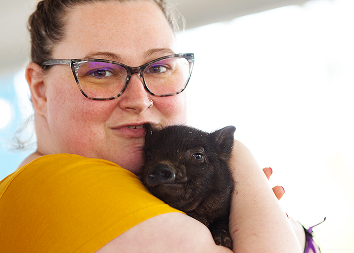 Woman with glasses holding a small black piglet close to her face inside a home.