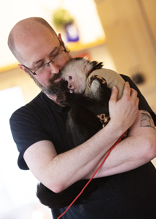A man with glasses and a beard holding a capuchin monkey close to his face, both appearing affectionate.