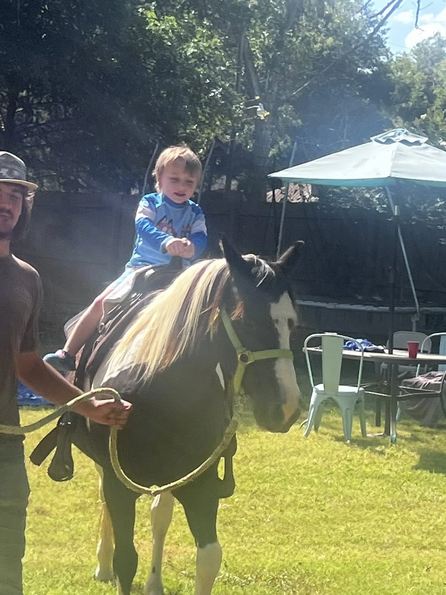 A young boy wearing a blue shirt riding a horse during an outdoor event on grass with tables, chairs, and a white canopy in the background.