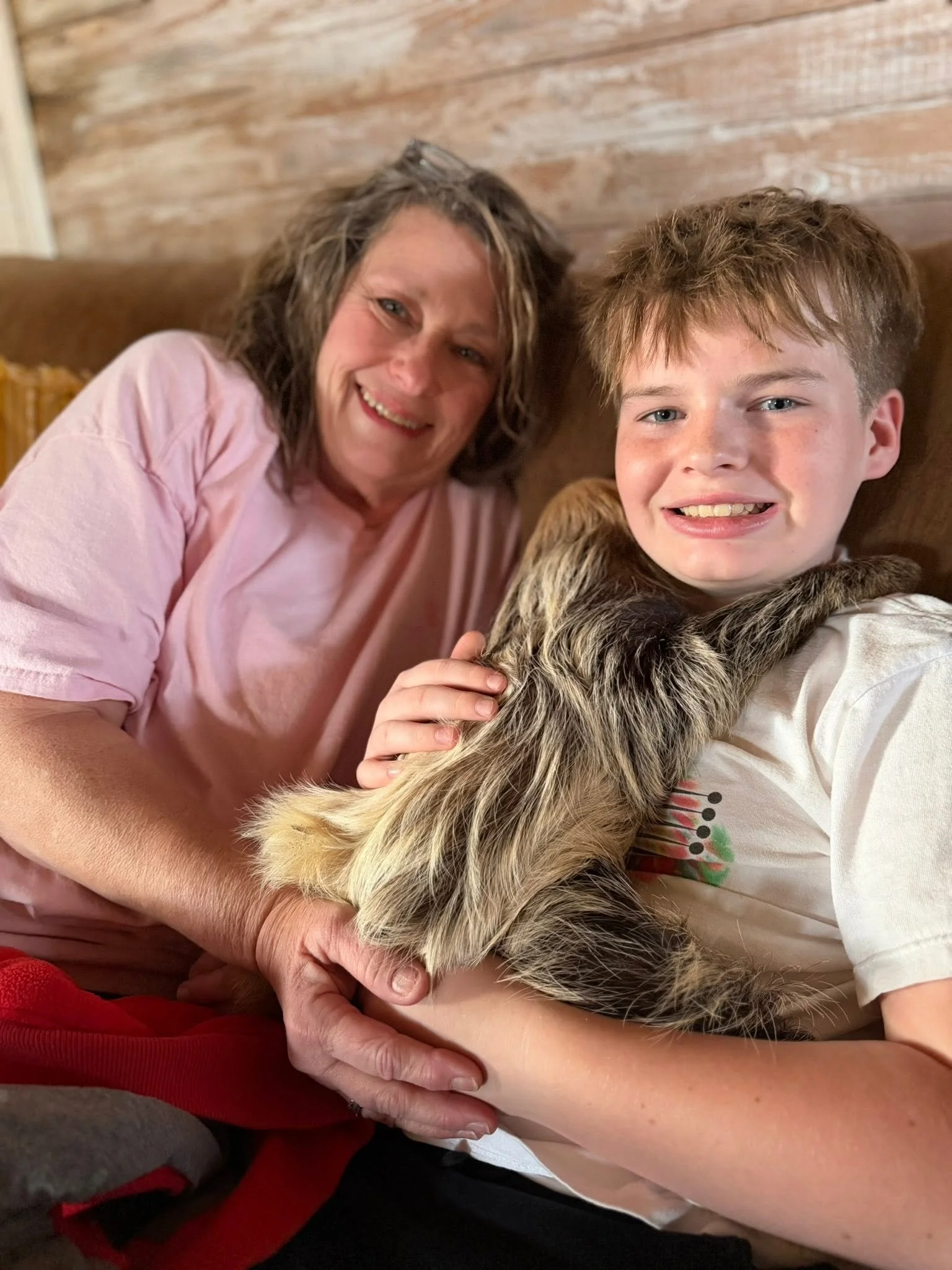 A woman and a boy sitting close together on a sofa, smiling at the camera, with the boy holding a dog with fur that has a mix of black, brown, and white colors.