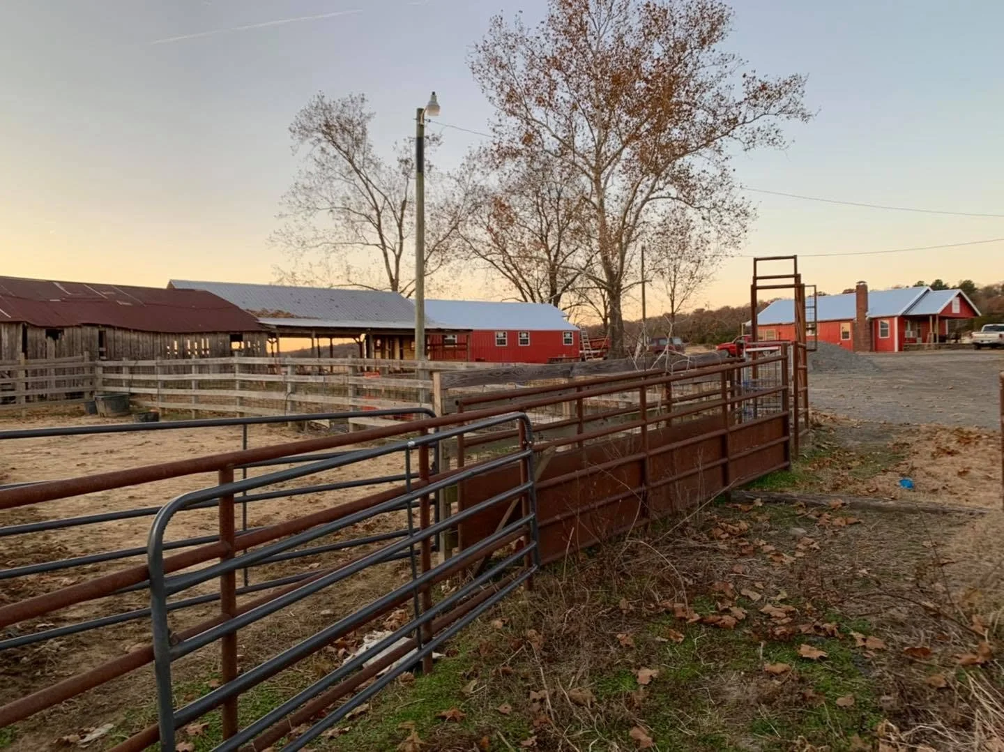 A farm scene at sunset with a fenced yard, red barn, trees, and a gravel driveway.