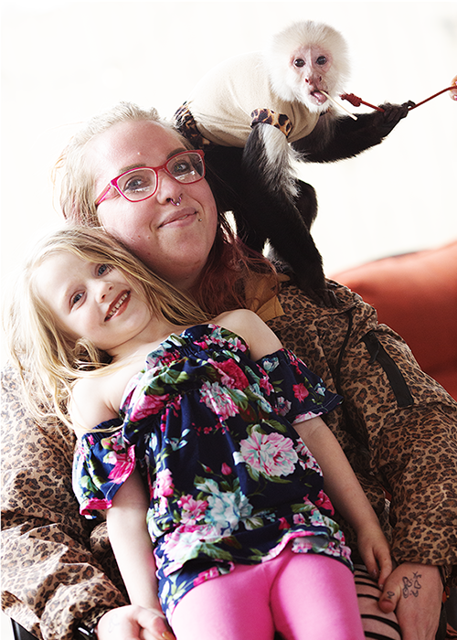 A woman with glasses and two young girls, one sitting on her lap and one perched on her shoulder, with a monkey on her shoulder holding a treat and tasting it.