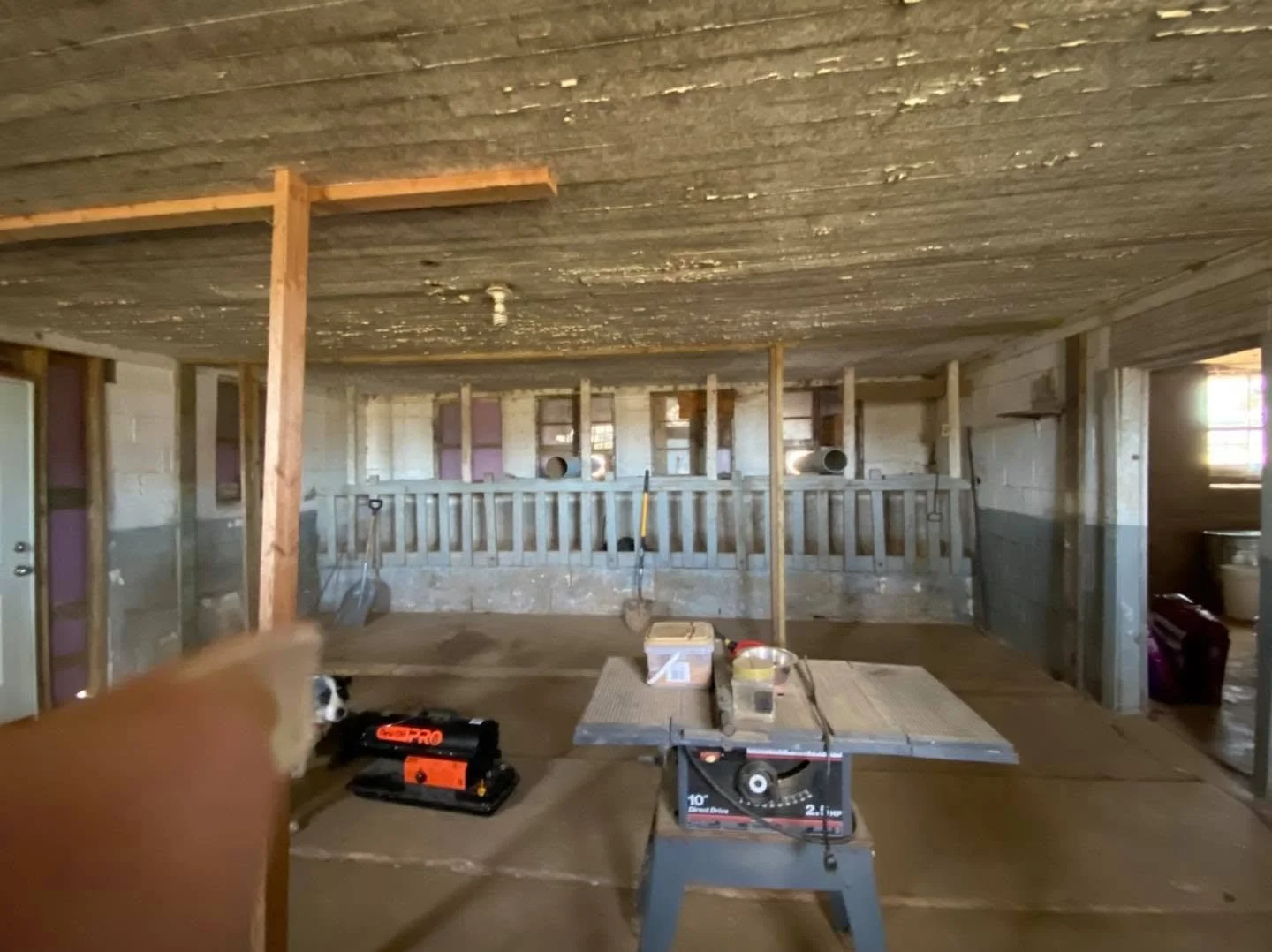An indoor construction site with unfinished walls, exposed wooden framing, and a plywood ceiling. There are construction tools and materials on the floor, including a miter saw, a cordless drill, a bucket, and other items. The space appears to be und