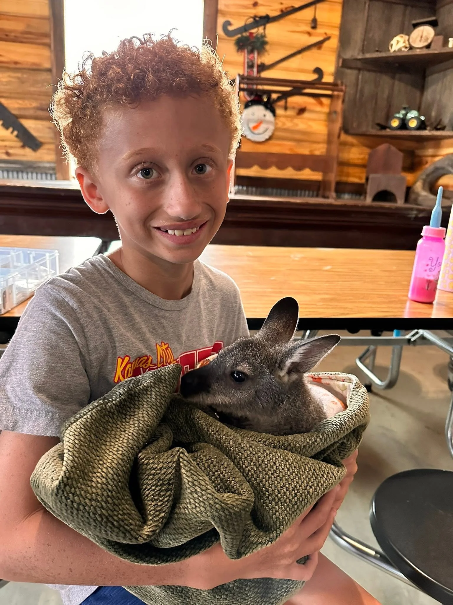 A young boy with curly red hair smiling while holding a small baby kangaroo in his arms, wrapped in a green blanket. The background shows wooden walls with rustic decorations and shelves.