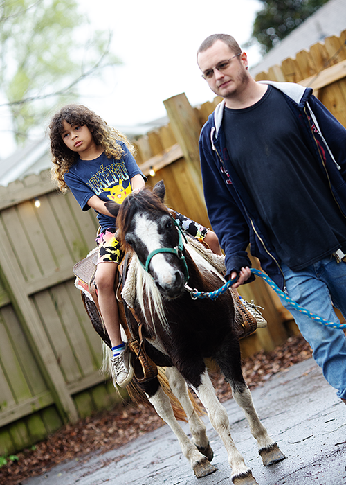 Young girl with curly hair riding a small pony, being led by a man outside near a wooden fence.