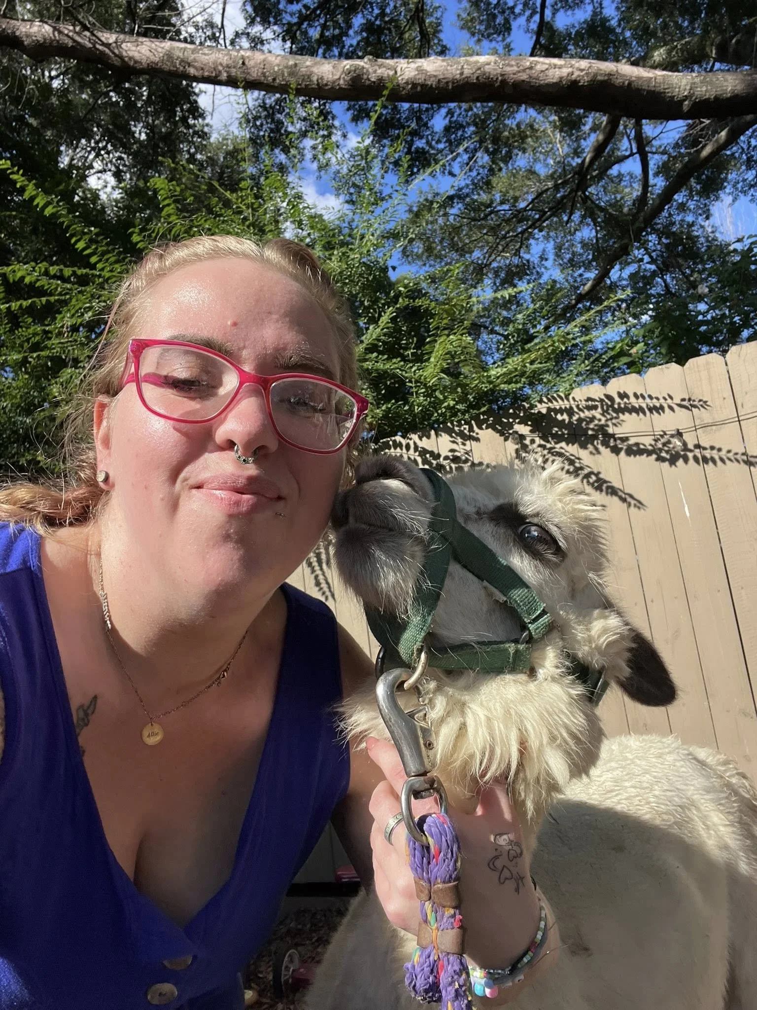 A woman with blonde hair, pink glasses, and tattoos on her arm is holding a black and white puppy outdoors in front of a wooden fence, trees, and blue sky.