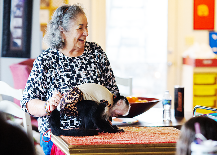 A woman with gray hair, wearing a black-and-white patterned top, is smiling while holding a small monkey dressed in a tan and leopard print outfit on a table.