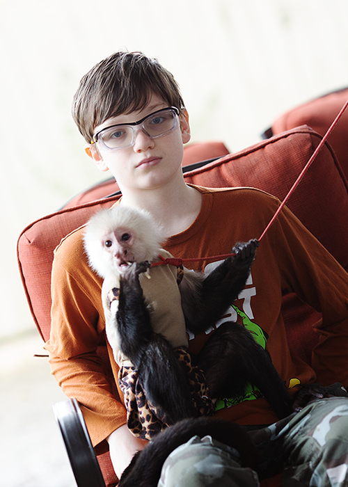 A young boy with glasses sitting on a red chair, holding a capuchin monkey on his lap, both looking at the camera.