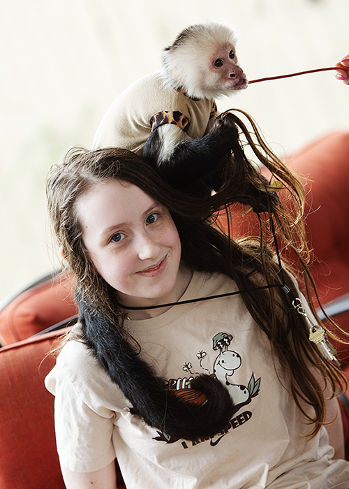 A girl with long brown hair smiling while a white and gray pet doll with a monkey face is sitting on her shoulder. A small monkey with light fur is on her head, reaching out with a stick.